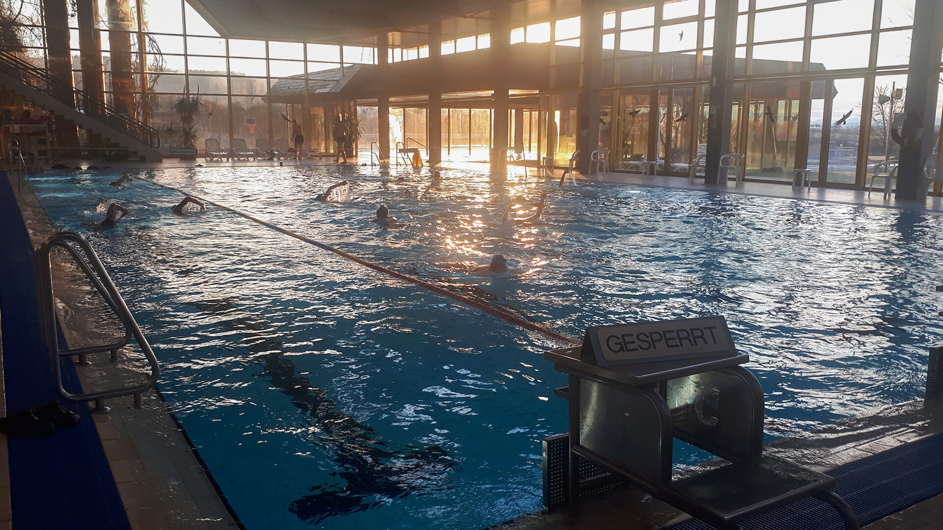 Hallen-Schwimmbecken mit mehreren Schwimmern, Startblock mit Gesperrt-Schild, verglaste Hallenwände