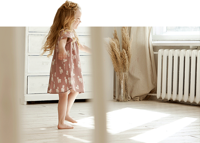 Mädchen im Kleid tanzt barfuß im Zimmer mit Holzfußboden, Kommode und Heizung
