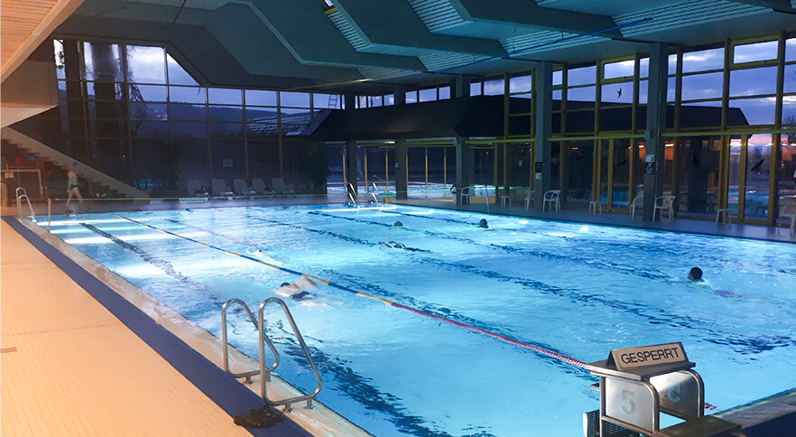 Hallen-Schwimmbecken mit mehreren Schwimmern, Startblock mit gesperrt-Schild und verglaste Hallenwänden in der Dämmerung
