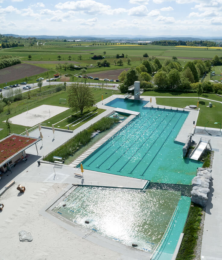 Luftaufnahme von Freibadanlage mit zwei Schwimmbecken, Tennisplatz, Grünanlage, Parkplatz, Wiesen und Felder im Hintergrund